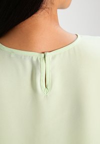 Mintgrön blus med rund halsringning, som har en nyckelhålsöppning på baksidan och en mjuk, slät textur.