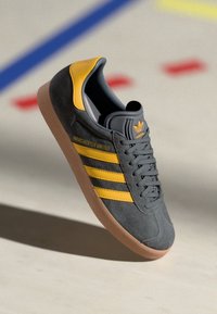 Gray suede sneaker with yellow stripes and logo. Features a padded collar, laces, and gum rubber sole. Embroidered branding on side.