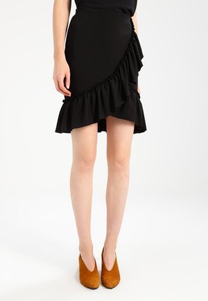 Zwarte asymmetrische rok met een gerimpelde zoom, gemaakt van gestructureerde stof. Gecombineerd met bruine suède enkellaarsjes met hoge hak.