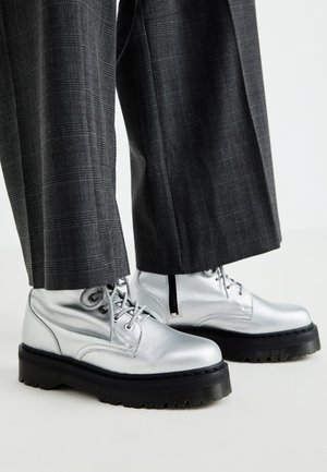 Bottes lacées argentées brillantes avec des semelles noires épaisses, portées avec un pantalon large à carreaux gris foncé.