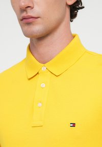 Gul polo skjorte laget av teksturert stoff med en tre-knapps knappestolpe. Merkverdig lite logo i rødt, hvitt og blått på venstre bryst.