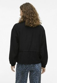 Čierna pulóvrová bunda s voľným strihom a rebrovanými manžetami, nosená s modrými nohavicami s leopardím vzorom. Tkanina má hladkú textúru.