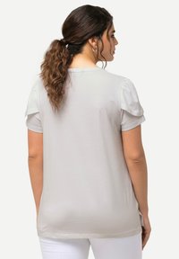 T-shirt grigio chiaro con maniche corte e vestibilità morbida. Tessuto soffice, scollo rotondo e volant sulle spalle aggiungono dettagli al design.