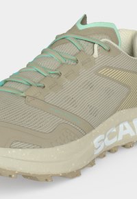 Chaussure de trail beige et vert clair avec dessus en maille texturée, lacets bleu clair et semelle robuste avec la marque "SCARPA".