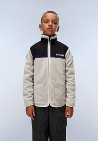 Fleecejacka i beiget och svart med hög krage, dragkedja, blå accentstickningar och två framfickor. Modellen bär cargobyxor.