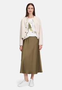 Hellbeige Cropped-Jacke, weißes T-Shirt mit Aufdruck, olivgrüner Satinsrock und weiße Sneakers. Einfaches, lässiges Outfit.