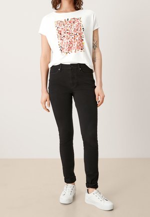 Camiseta blanca con un patrón abstracto en rosa y amarillo, combinada con jeans ajustados negros. Zapatillas blancas de diseño simple y con los extremos de los jeans enrollados.