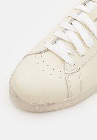 White leather sneaker with a smooth texture, rounded toe, and perforated details. Features flat white laces and a slightly worn rubber sole.