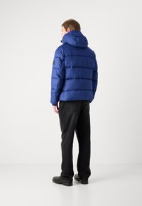 Mėlynas pūkinis švarkas su horizontaliais quilted skyriumi ir gobtuvu, pagamintas iš blizgaus medžiagos. derinamas su juodomis kelnėmis ir batais.