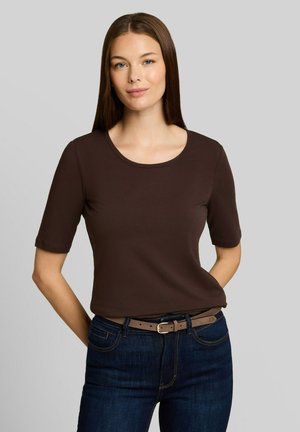 Frau mit glatten braunen Haaren, die ein eng anliegendes braunes Kurzarmoberteil trägt, das in dunkelblaue Jeans mit braunem Gürtel gesteckt ist, stehend vor einem grauen Hintergrund.