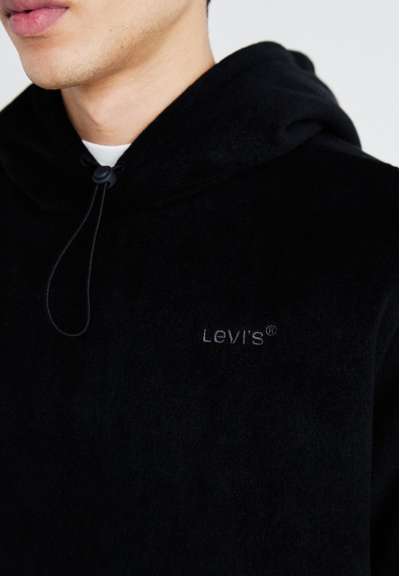 Haut en polaire noir avec un cordon de serrage réglable, présentant un logo Levi's brodé discret sur la poitrine. Texture douce, coupe décontractée.