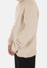 Beżowy dzianinowy sweter z golfem z prążkowanym wykończeniem u dołu i na mankietach, o fakturowanej powierzchni. Nałożony na czarne spodnie, widoczny z boku.