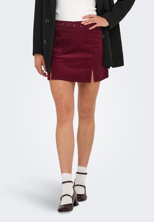 Femme portant une jupe mini bordeaux avec des fentes à l'avant, des chaussettes blanches, des escarpins Mary Jane marron foncé, un blazer noir et un haut blanc.
