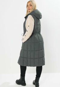 Gilet matelassé gris avec capuche, présentant des manches en tricot beige et des accents de boutons. Le design comprend une taille élastique et une longueur coupe-vent.