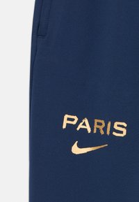 Marina blå sweatpants med en slät yta; har "PARIS" och Nike-logotyp i metallic guldtryck på vänster lår.