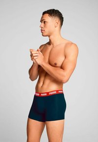 Black boxer shorts with a red Puma logo waistband, fitted design, and soft material, shown on a male model from the side.