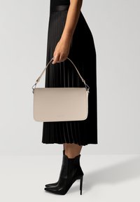 Bolso de piel texturizada beige, con forma rectangular y correa desmontable, sostenido por una persona que lleva una falda negra con pliegues y botines negros.