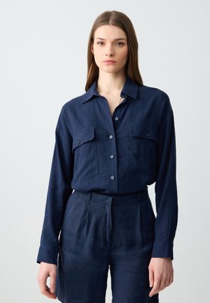 Donna con lunghi capelli castani che indossa una camicia button-up blu scuro con tasche frontali e pantaloni a vita alta coordinati, in piedi di fronte a uno sfondo neutro.