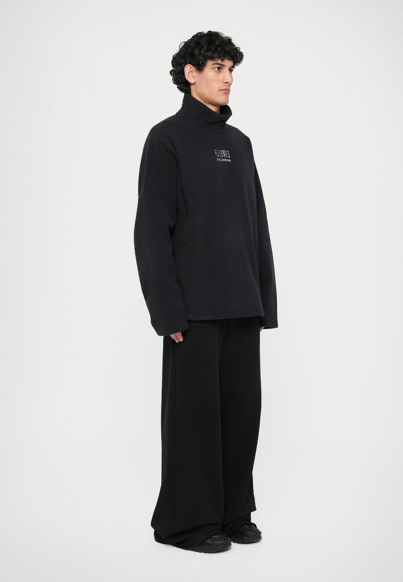 Black oversized pullover with high collar, made of soft fabric. Features front logo and loose-fitting black trousers. Footwear: black sneakers.