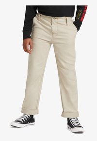 Beige Baumwollhosen mit geradem Bein, ausgestattet mit einem Knopfverschluss, Gürtelschlaufen und aufgerollten Bündchen, kombiniert mit schwarzen High-Top-Sneakern.