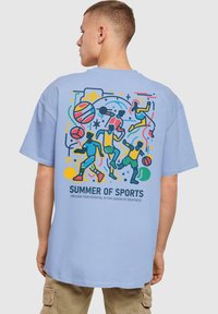 Světle modré tričko s barevnou grafikou sportovců, basketbalů a nápisem "LETNÍ SPORTY" v černé barvě, s hravými designovými prvky.