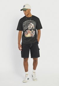 Mann trägt schwarzes T-Shirt mit Chicago-Grafik, schwarze Shorts, weiße Turnschuhe und eine weiße Baseballkappe mit grünem Schirm, steht vor weißem Hintergrund.