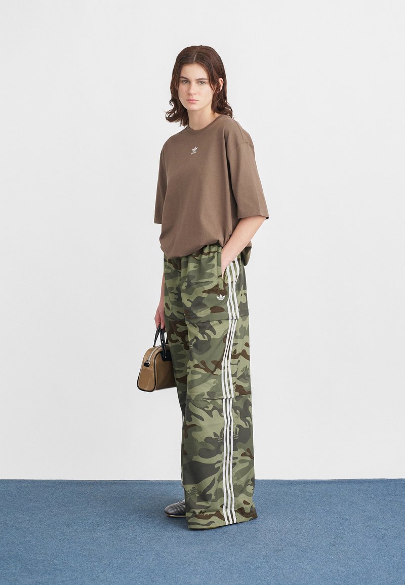 Braunes oversized T-Shirt, camouflierte weite Hose mit weißen Streifen und eine kleine schwarze Handtasche. Die Umgebung hat einen blauen Teppich und eine weiße Wand.