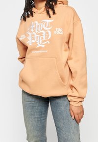 Sudadera con capucha de suave tela melocotón con un bolsillo delantero. Presenta un atrevido estampado de texto blanco, un corte relajado y puños acanalados.