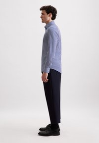 Camicia a righe blu e bianche con colletto, abbinata a pantaloni sartoriali scuri e scarpe nere, mostrata di lato.
