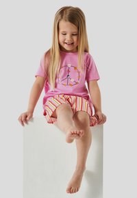 T-shirt en coton rose avec un motif de signe de paix et des accents floraux, associé à un short rayé dans des tons de rose, orange et blanc. Assis sur une surface blanche.