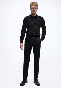 Camicia elegante nera con motivo a fantasia e maniche lunghe, abbinata a pantaloni scuri e scarpe nere stringate. Presenta un design aderente e una texture liscia.