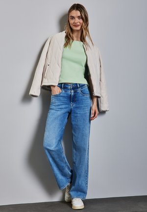 Lys beige quiltet bomberjakke over en mintgrøn top, parret med løse blå jeans og hvide sneakers, mod en grå baggrund.