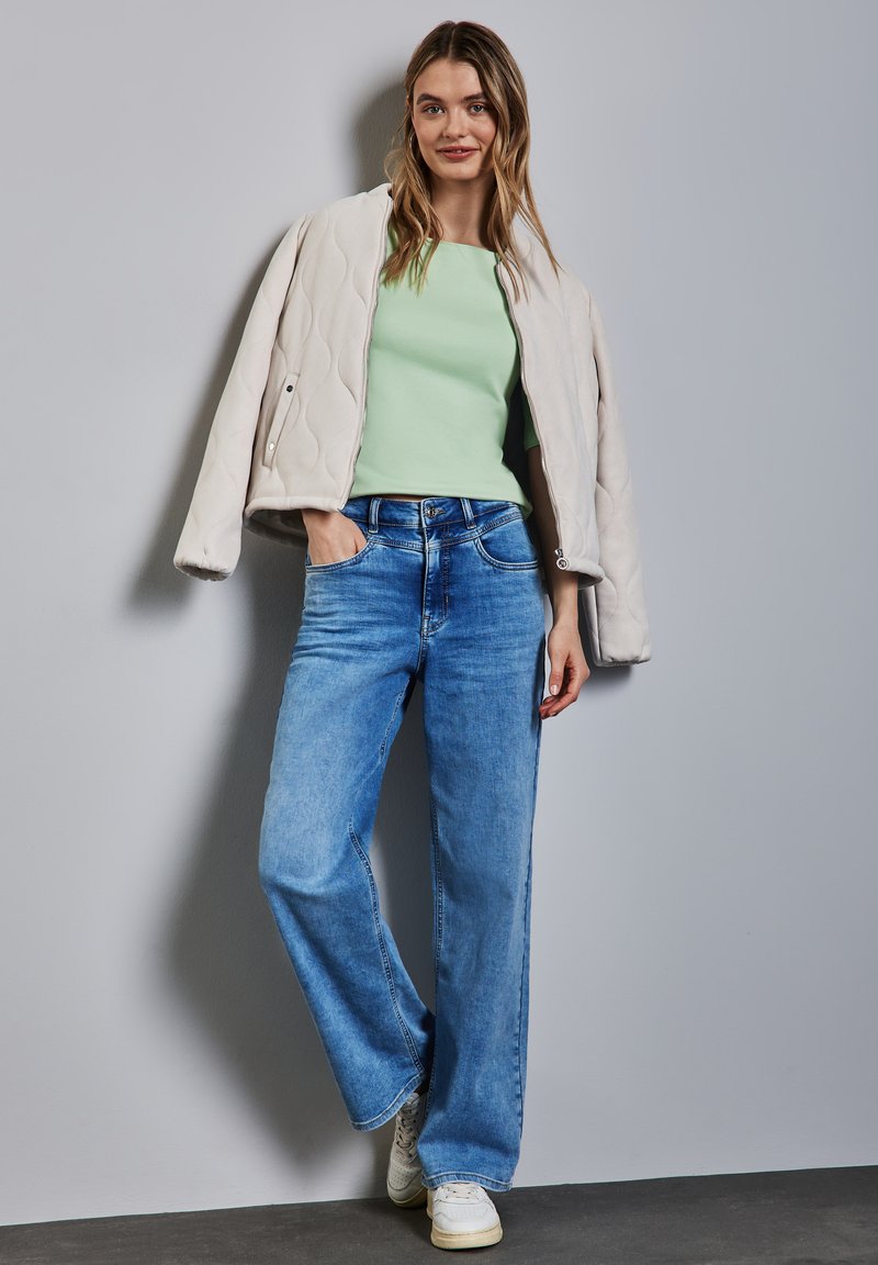 Giubbotto bomber trapuntato beige chiaro sopra una maglietta verde menta, abbinato a jeans blu larghi e sneakers bianche, su uno sfondo grigio.