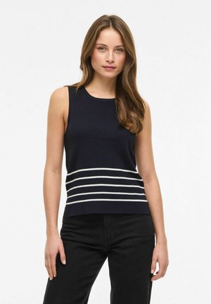 Ung kvinde med brunt hår iført en ærmeløs navyblå top med fire hvide horisontale striber og sorte bukser mod en hvid baggrund.