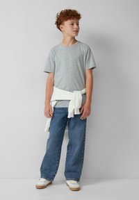 Junge mit lockigem roten Haar, der ein graues T-Shirt, blaue Jeans, weiße Turnschuhe und einen um die Hüfte gebundenen weißen Pullover trägt, steht vor einer schlichten Wand.