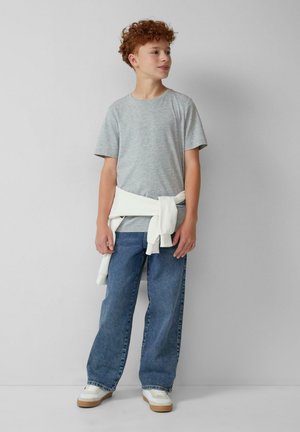 Junge mit lockigem roten Haar, der ein graues T-Shirt, blaue Jeans, weiße Turnschuhe und einen um die Hüfte gebundenen weißen Pullover trägt, steht vor einer schlichten Wand.