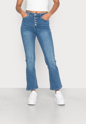Jeans de cintura alta e boca de sino em denim azul claro, com fecho de botão, bolsos frontais e bainha limpa. Combinados com ténis brancos de plataforma.