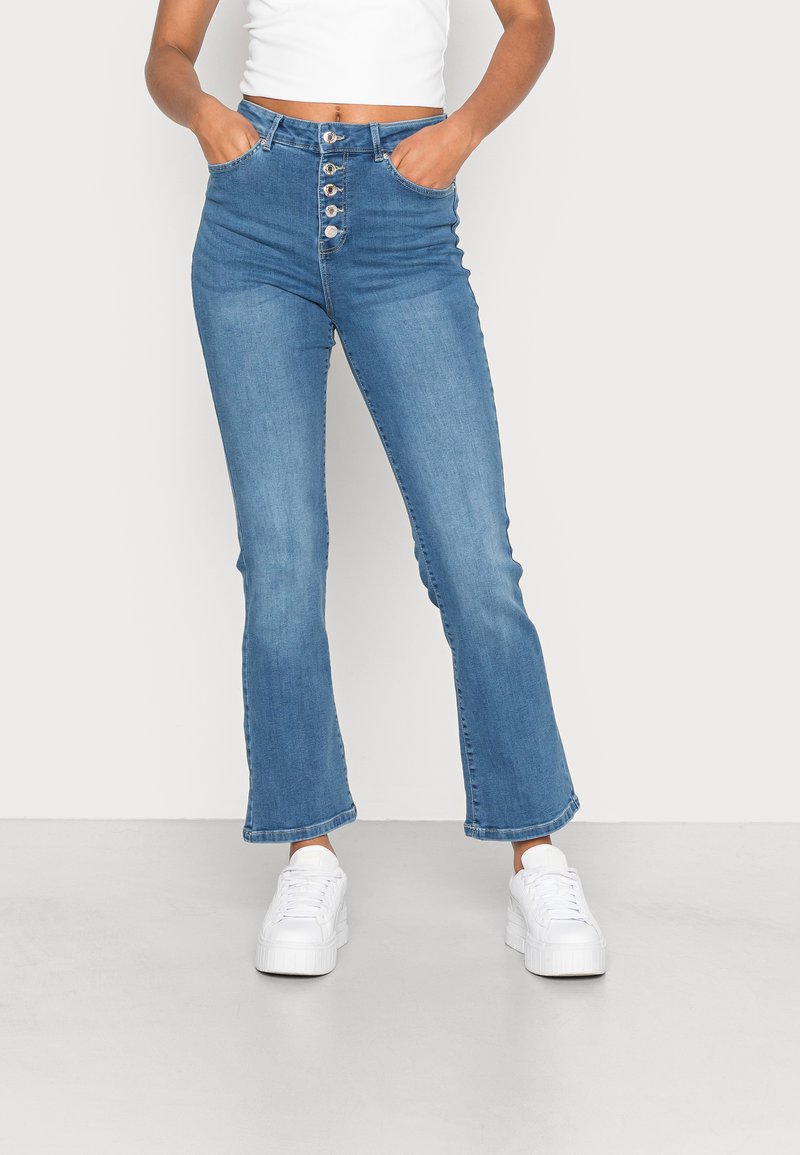 Jeans de cintura alta e boca de sino em denim azul claro, com fecho de botão, bolsos frontais e bainha limpa. Combinados com ténis brancos de plataforma.