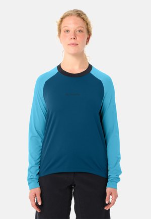 Vrouw met blond haar draagt een blauw-groen sportshirt met lange mouwen en zwarte broek, staand tegen een effen grijze achtergrond.