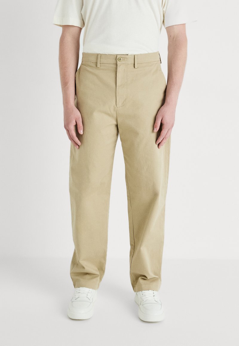 Pantaloni din bumbac bej cu un design drept, având închidere frontală cu buton și buzunare laterale. Purtați cu adidași albi.