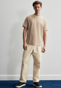 Beige bomulds T-shirt parret med lyse beige bukser og sorte sneakers. Outfittet har en afslappet pasform og en blid tekstur.