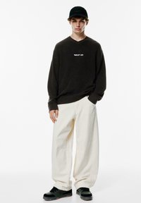Pulover tricotat cu coaste de culoare maro închis, având imprimate "SIMPLIFY LIFE", purtat cu pantaloni crema supradimensionați și adidași negri cu accente verzi. Șapcă neagră.