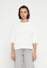 Λευκό πλεκτό πουλόβερ με στρογγυλή λαιμόκοψη, μανίκια ράγκλαν τριών τετάρτων και άνετη εφαρμογή, σε συνδυασμό με ανοιχτογκρι τζιν.