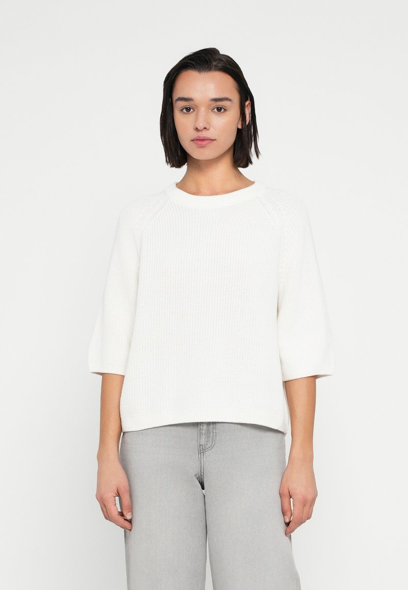 Λευκό πλεκτό πουλόβερ με στρογγυλή λαιμόκοψη, μανίκια ράγκλαν τριών τετάρτων και άνετη εφαρμογή, σε συνδυασμό με ανοιχτογκρι τζιν.