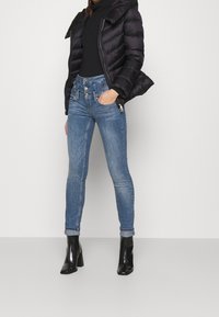 Sort pufferjakke med hætte over en tætsiddende sort turtleneck, parret med lyseblå skinny jeans med høj talje og knapdetaljer. Sorte hæle støvler.