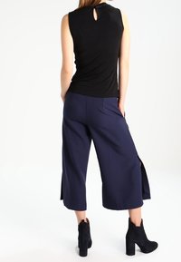 Haut noir sans manches avec une ouverture en forme de trou de serrure dans le dos, associé à un pantalon culotte bleu marine à fentes latérales, et des bottines noires à talon épais.