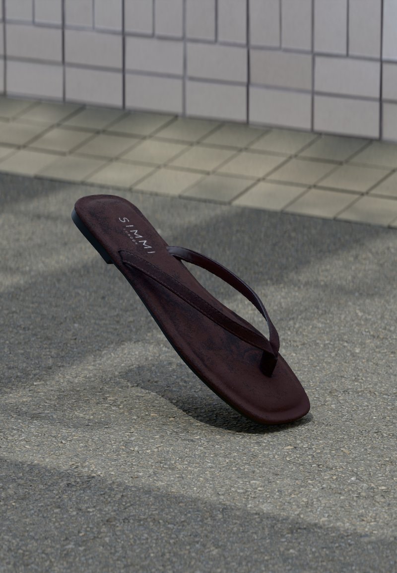 Ein einzelner dunkelbrauner Flip-Flop steht aufrecht auf einer gepflasterten Oberfläche vor einer hellfarbenen, gefliesten Wand.