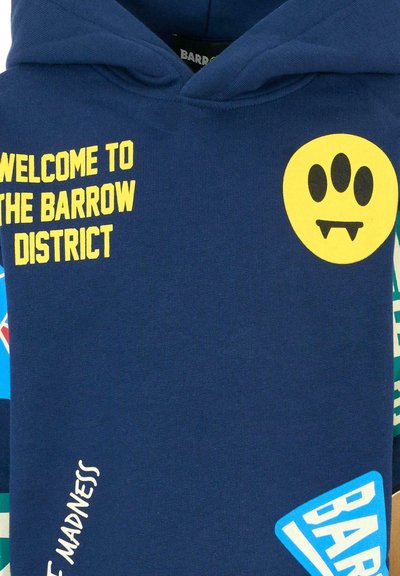 Felpa blu navy con testo giallo "BENVENUTO NEL DISTRETTO DI BARROW", una grafica gialla con un volto e varie toppe colorate sul davanti.