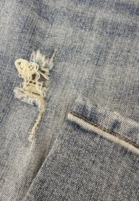 Lichtblauwe denimstof met gescheurde rafeldetails, contrasterende donkere stiksels langs de randen en een klein zakje rechts onderaan.