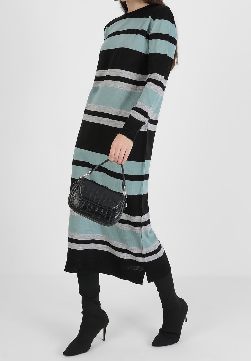 Robe en maille rayée avec des rayures noires, turquoise et grises ; manches longues. Sac à main noir texturé tenu à la main. Coupe longueur cheville avec manches ajustées.
